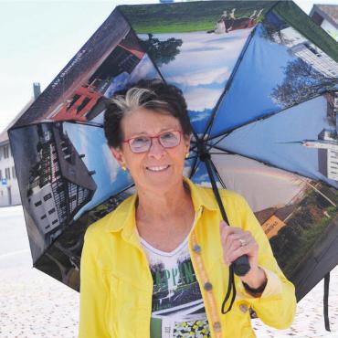 Trocken durch das Dorf - Frau Gemeindeammann Rosmarie Groux zeigt, dass der Regenschirm auch als Schattenspender bei sonnigem Wetter eingesetzt werden kann. Bild: Roger Wetli